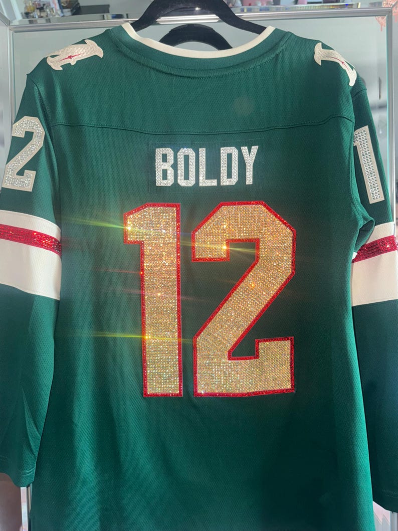 Puede incluir: Una camiseta verde con el nombre "BOLDY" y el n&uacute;mero "12" en adornos brillantes. La camiseta tiene detalles blancos y rojos en las mangas y los hombros. El n&uacute;mero "2" tambi&eacute;n es visible en el hombro izquierdo.