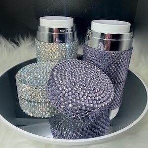 May include: Two cylindrical containers with silver metallic rims and white tops, each fully encrusted with small sparkling rhinestones. One container is covered in clear iridescent crystals, while the other is covered in light purple crystals.