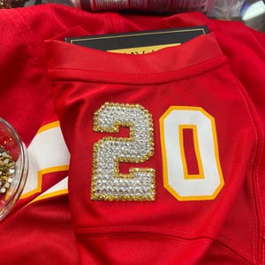 Puede incluir: Camiseta roja con el n&uacute;mero 20 en blanco con contorno dorado. El n&uacute;mero est&aacute; adornado con pedrer&iacute;a transparente y dorada. La camiseta tiene una raya blanca y dorada en el hombro.