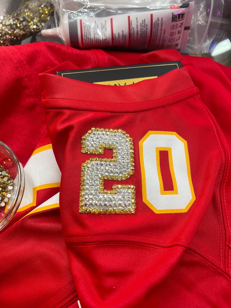 Puede incluir: Camiseta roja con el n&uacute;mero 20 en blanco con contorno dorado. El n&uacute;mero est&aacute; adornado con pedrer&iacute;a transparente y dorada. La camiseta tiene una raya blanca y dorada en el hombro.