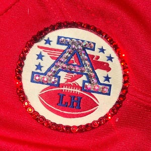 Puede incluir: Un parche circular con un borde de pedrer&iacute;a roja. El parche presenta un fondo blanco con una letra "A" azul y pedrer&iacute;a, estrellas azules y un &aacute;guila roja. Un bal&oacute;n de f&uacute;tbol rojo con "LH" est&aacute; en la parte inferior.