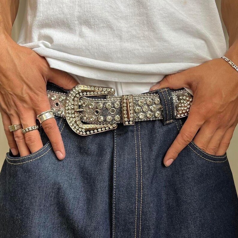 Puede incluir: Un cintur&oacute;n plateado con una hebilla grande y adornada y tachonada con pedrer&iacute;a transparente. El cintur&oacute;n se usa con jeans de mezclilla azul oscuro. La hebilla es rectangular y tiene un dise&ntilde;o detallado.