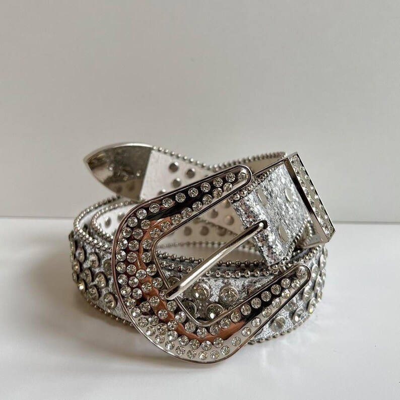 Puede incluir: Un cintur&oacute;n plateado y blanco adornado con brillantes pedrer&iacute;a. El cintur&oacute;n presenta una hebilla plateada grande y est&aacute; cubierto de peque&ntilde;os brillantes redondos. El cintur&oacute;n est&aacute; hecho de cuero blanco y tiene tachuelas plateadas en los bordes. Un accesorio de moda.