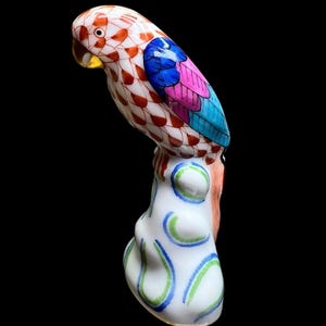 May include: A ceramic parrot figurine with a white body and a colorful design. The parrot's head is white with red and orange accents, and its wings are blue, pink, and turquoise. The base is white with blue and green swirls.
