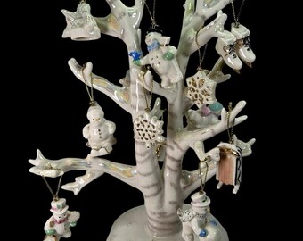 May include: A ceramic Christmas tree with various ornaments, including snowmen, ice skates, and an angel. The tree is white with gold accents and sits on a round base. The ornaments are suspended by gold strings.
