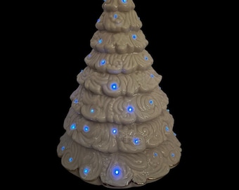 May include: A ceramic Christmas tree with a cream-colored glaze and blue lights. The tree has a bow on top and gold trim. The lights are evenly spaced throughout the tree, creating a festive glow.