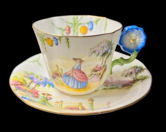 May include: A decorative teacup and saucer set with a blue flower handle. The cup and saucer are decorated with a painted landscape scene and gold trim. The set is designed with a vintage aesthetic and is ideal for tea.