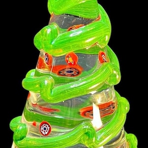 May include: A clear glass Christmas tree with green swirls and colorful decorative accents. The tree is approximately 6 inches tall and has a rounded base. The design includes red, orange, and blue details.