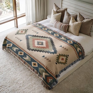 Pode incluir: Uma manta tecida com um padrão geométrico em tons de bege, verde sálvia, azul escuro e terracota. A manta tem uma borda com franjas e está colocada sobre uma cama com várias almofadas decorativas em tons neutros de creme e castanho.