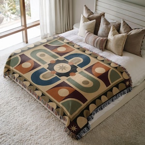 May include: A woven throw blanket featuring a geometric pattern of arches, circles, and a central starburst motif in shades of sage green, terracotta, navy blue, and cream. The blanket has a fringed edge and is draped over a bed with several pillows.