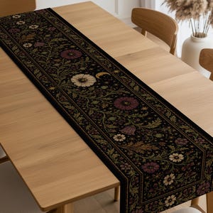 May include: A long, rectangular black table runner featuring a detailed botanical print with cream, burgundy, and olive green flowers, winding vines, and small golden celestial motifs including stars and crescent moons arranged in a repeating pattern.