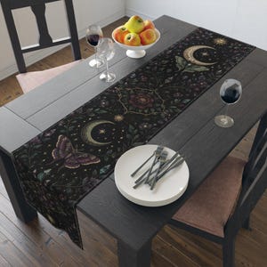 May include: A dark table runner with a celestial design featuring moons, stars, and butterflies. The runner is placed on a dark wooden table set with plates, silverware, and wine glasses. A bowl of apples sits in the center.
