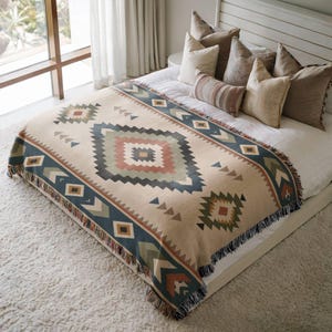 May include: A woven throw blanket featuring a geometric pattern in shades of beige, sage green, dark blue, and terracotta. The blanket has a fringed edge and is draped over a bed with several decorative pillows in neutral tones of cream and brown.