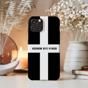 May include: Black and white phone case featuring a cross design with the text "KERNOW BYS VYKEN" in white. The case is on a white surface with a blurred background of flowers and a candle.