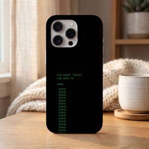 Pode incluir: Capa de telefone preta com texto verde exibindo um programa BASIC simples que imprime "IYKYK" repetidamente. A capa tem um acabamento brilhante e um recorte para a câmera.
