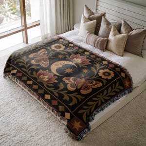May include: A woven throw blanket featuring a black background with a central crescent moon, stars, two large butterflies, and floral motifs in shades of gold, brown, and muted red. The blanket has a decorative diamond border and fringed edges.
