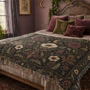 May include: A woven throw blanket featuring a dark background with a botanical pattern of cream, burgundy, and green flowers, leaves, and small golden crescent moons. The rectangular blanket has a decorative border and frayed edges on the sides.