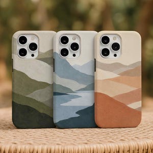 May include: Three phone cases with landscape designs. The cases feature a mountain range in shades of green, blue, and orange. Each case has a camera cutout and a neutral-colored border.