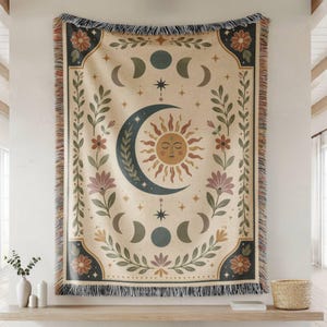 May include: A woven throw blanket featuring a central sun and crescent moon face design surrounded by moon phases, stars, and floral botanical motifs in muted tones of cream, dark blue, terracotta, and sage green with a fringed edge finish.
