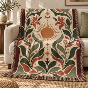 May include: A woven throw blanket featuring a central sun motif, crescent moon, and star icons. The design includes symmetrical botanical patterns in shades of forest green, terracotta, and cream, finished with a fringed edge on the bottom.