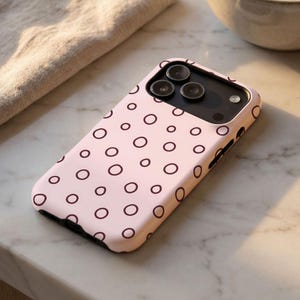 Può includere: Una custodia per smartphone rosa chiaro con un motivo ripetuto di piccoli cerchi marrone scuro. La custodia presenta un ritaglio rettangolare per il modulo fotocamera, che contiene tre lenti circolari e un flash, e l'accesso ai tasti laterali.