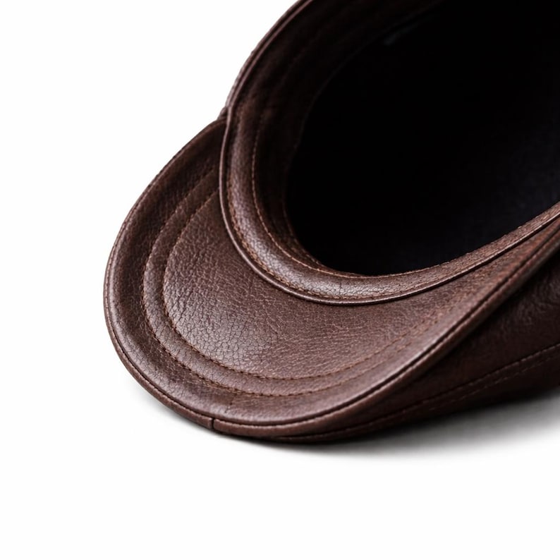 May include: Close-up of a brown leather newsboy cap. The cap features a curved brim and a stitched design. The interior of the cap is black. The cap is made of a smooth, textured leather material.