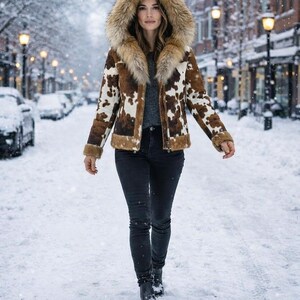 May include: A woman wearing a brown and white cowhide print jacket with a fur-lined hood. The jacket has a zipper closure and brown trim. She is wearing black jeans and boots, walking on a snow-covered street.