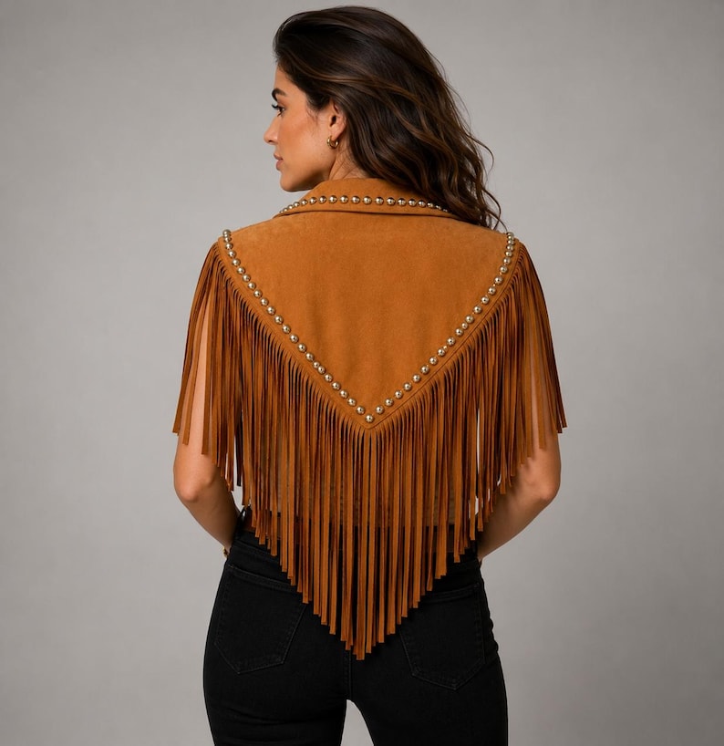 May include: A tan suede vest featuring a V-shaped back yoke adorned with metallic studs. The garment has long, flowing fringe detailing that extends from the shoulders down to a point at the center back, creating a layered appearance.