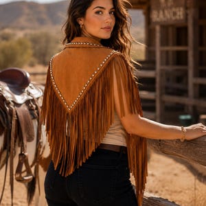 May include: A tan suede vest featuring a V-shaped back panel adorned with silver-toned metal studs along the edges and long, flowing fringe detailing that extends from the shoulders down to the lower back area.