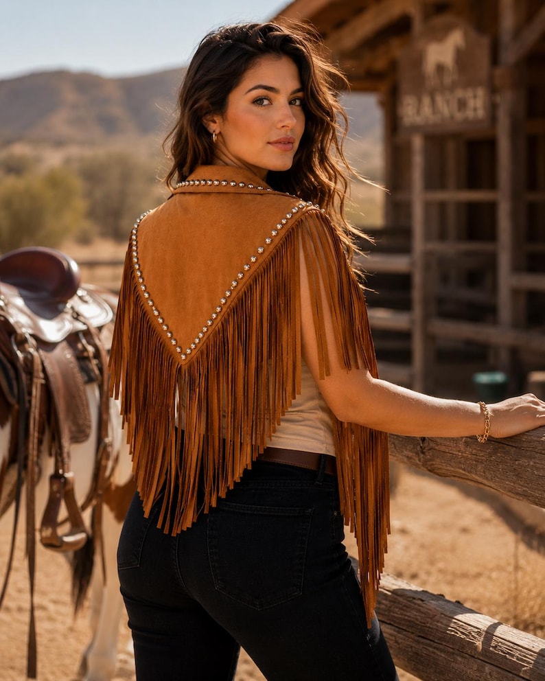 May include: A tan suede vest featuring a V-shaped back panel adorned with silver-toned metal studs along the edges and long, flowing fringe detailing that extends from the shoulders down to the lower back area.
