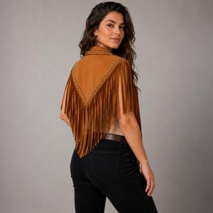 May include: A tan suede vest featuring a V-shaped back panel adorned with metallic studs along the edges and long, flowing fringe detailing that extends from the shoulders down to the waist, paired with black denim jeans and a brown belt.