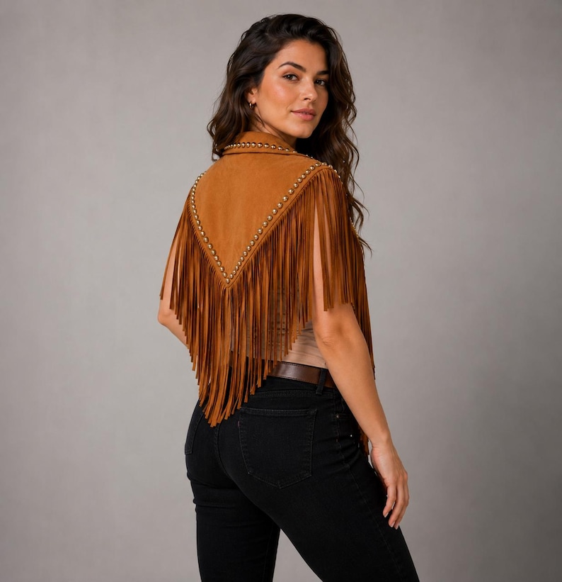 May include: A tan suede vest featuring a V-shaped back panel adorned with metallic studs along the edges and long, flowing fringe detailing that extends from the shoulders down to the waist, paired with black denim jeans and a brown belt.
