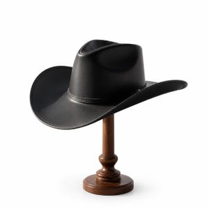 Könnte beinhalten: Ein schwarzer Cowboyhut mit breiter Krempe und Zierband, präsentiert auf einem Hutständer aus dunklem Holz. Der Hut hat eine klassische Form und ist aus glattem Material gefertigt.