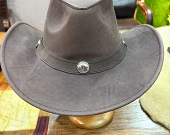 May include: A brown cowboy hat with a wide brim and a pinched crown. The hat features decorative silver-coloured metal accents and a leather band. The hat is made of a suede-like material and is displayed on a wooden stand.