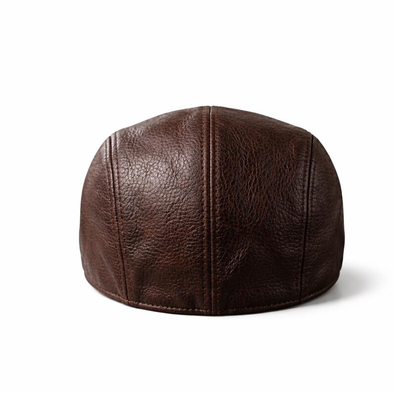 May include: A brown leather newsboy cap. The cap has a rounded crown and a short, stiff brim. The leather has a textured appearance with visible stitching details. The cap is a solid brown color and appears to be new.