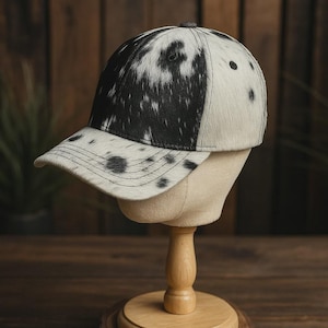 May include: A baseball cap with a cowhide pattern. The cap features a black and white spotted design on the crown and brim, with a solid black button on top. The cap is displayed on a wooden mannequin head.