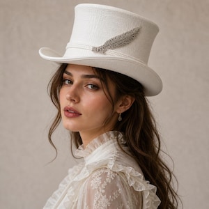 May include: A cream-coloured top hat with a textured finish and a silver feather embellishment. The hat features a broad brim and a band around the crown. The feather is fixed to the side.