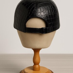 May include: Black baseball cap with a textured, crocodile-like pattern. The cap features a curved brim, a button on top, and an adjustable strap with a buckle. The cap is displayed on a mannequin head.