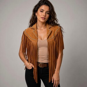 May include: A tan suede vest featuring long fringe detailing along the front panels and shoulders. The garment is accented with small, metallic silver studs lining the lapels and shoulder seams, creating a structured, sleeveless silhouette.