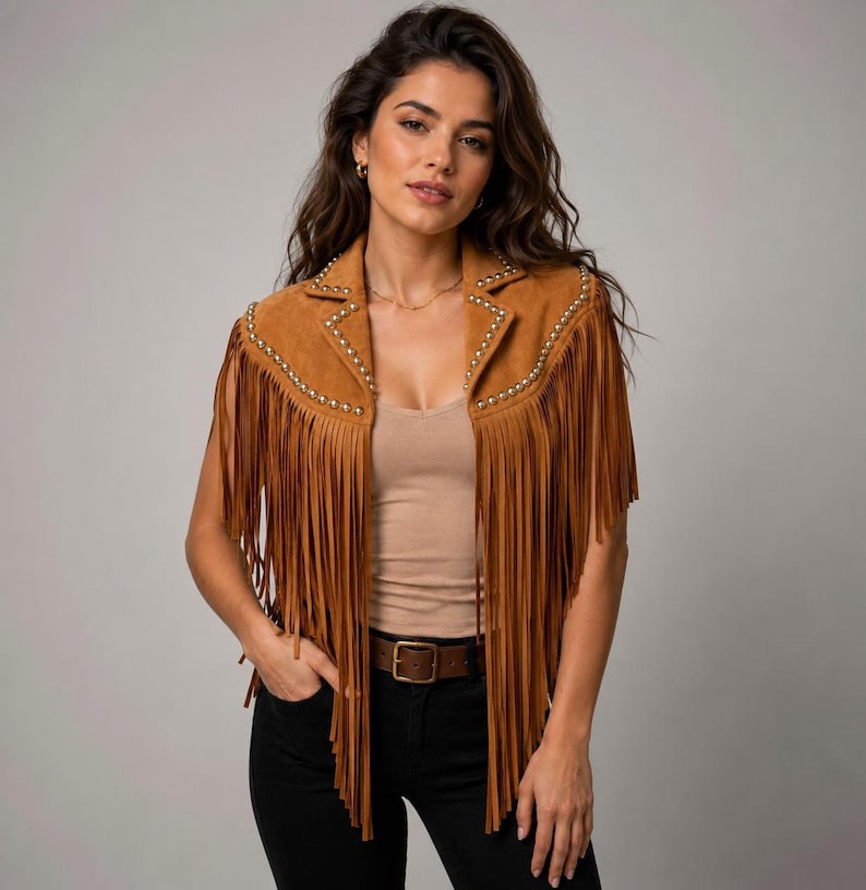 May include: A tan suede vest featuring long fringe detailing along the front panels and shoulders. The garment is accented with small, metallic silver studs lining the lapels and shoulder seams, creating a structured, sleeveless silhouette.