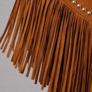 May include: A tan suede garment featuring long, thin fringe strips hanging vertically. A diagonal seam runs across the upper portion, accented by a single row of small, polished silver-toned metal studs spaced evenly along the stitched line.