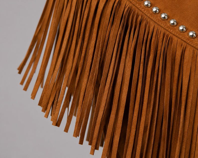 May include: A tan suede garment featuring long, thin fringe strips hanging vertically. A diagonal seam runs across the upper portion, accented by a single row of small, polished silver-toned metal studs spaced evenly along the stitched line.