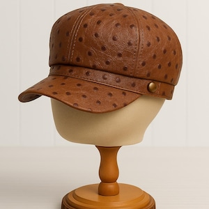 May include: A brown leather newsboy cap with a rounded crown and a short, curved brim. The cap features a pattern of small, evenly spaced dark brown dots. It is displayed on a mannequin head with a wooden stand.