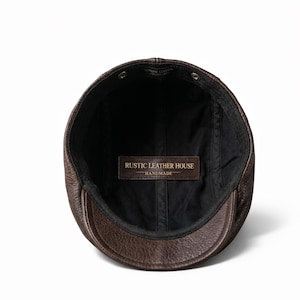 May include: A brown leather cap with a short brim, viewed from the inside. The cap has a black lining and a leather tag that reads "RUSTIC LEATHER HOUSE - HANDMADE". The cap is made of genuine leather.