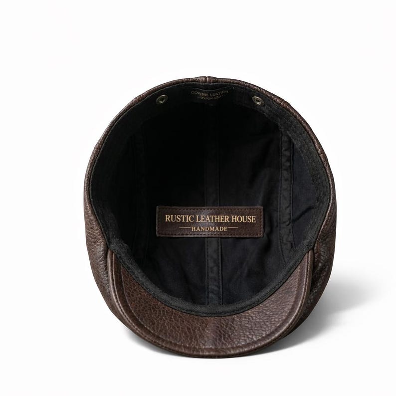 May include: A brown leather cap with a short brim, viewed from the inside. The cap has a black lining and a leather tag that reads "RUSTIC LEATHER HOUSE - HANDMADE". The cap is made of genuine leather.