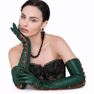 Könnte beinhalten: Grüne Leder-Opernhandschuhe mit roter Paspelierung und Knopfdetails. Die Handschuhe sind lang und reichen über die Ellbogen hinaus. Das Model trägt außerdem eine schwarze Perlenkette und Ohrringe.