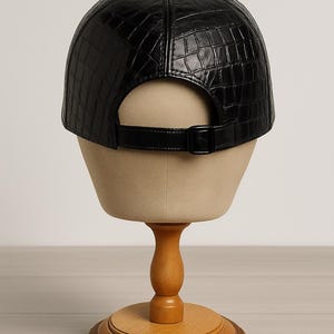 May include: Black baseball cap with a textured, crocodile-like pattern. The cap features a curved brim, a button on top, and an adjustable strap with a buckle. The cap is displayed on a mannequin head.