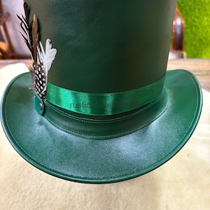 May include: A dark green top hat with a wide brim and a green ribbon band. The hat is adorned with a cluster of feathers in black, white, and brown. The text "rustiche" is visible on the ribbon.