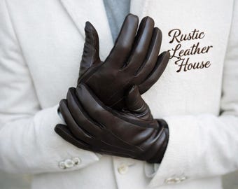 May include: Dark brown leather gloves. The gloves are smooth and form-fitting, with visible stitching. The gloves are worn over a white coat. The text "Rustic Leather House" is printed on the coat.