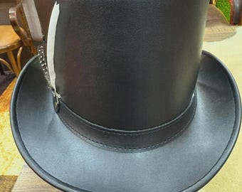 Op de afbeelding: Een zwarte hoge hoed met een brede rand en een decoratieve veer. De hoed is gemaakt van een glad, donker materiaal en heeft een band rond de kroon. De veer is wit met donkere accenten.